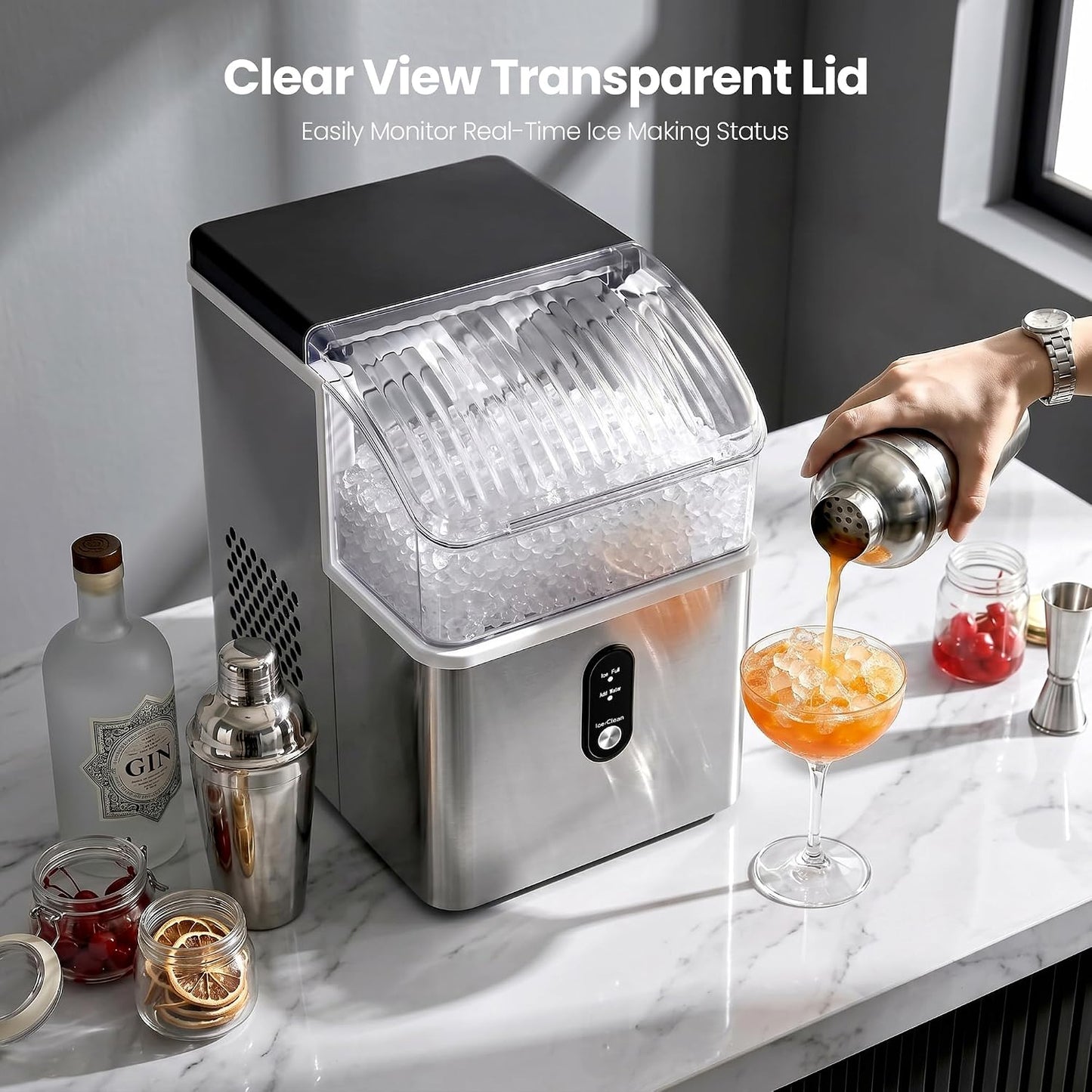 A countertop ice machine with a clear view transparent lid, shown on a white marble kitchen counter with a hand pouring an orange cocktail into a glass. The machine is filled with crushed ice, and bar accessories like a shaker, bottles, and jars are arranged around it.